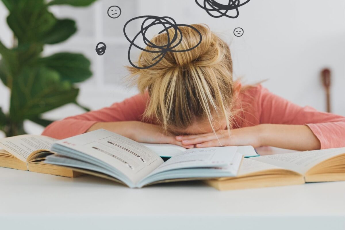 A person with blonde hair in a messy bun is slumped over a desk, their face buried in their arms amidst several open books. Above their head are illustrated squiggly lines and unhappy face emojis, symbolizing stress or confusion