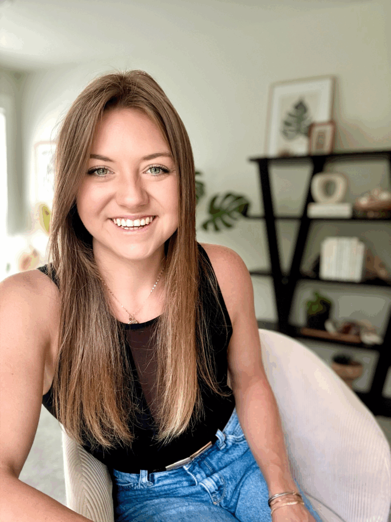 Jordan Casey of wellness IQ sits in a white chair, wearing blue jeans and a black top. in the background are black shelves and a leafy green plant.