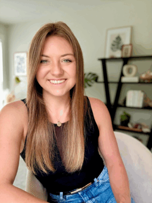 Jordan Casey of wellness IQ sits in a white chair, wearing blue jeans and a black top. in the background are black shelves and a leafy green plant.